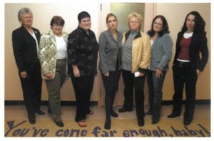 Fall 2006 photo of seven gender-equality leaders, standing with a banner reading "You've come far enough, baby!"